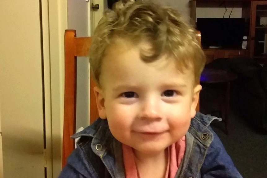 Two-year-old Connor Horan sits at a table, looking at the camera smiling.