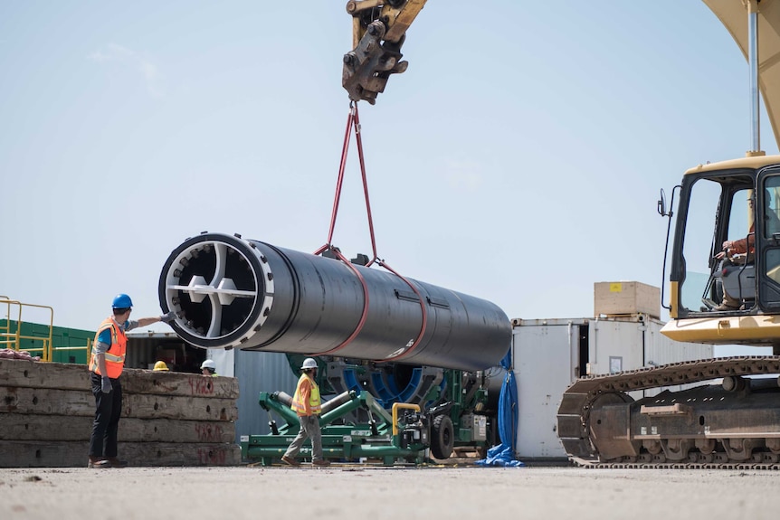 A crane lifts a large section of plastic pipe as men in high vision gear watch on.