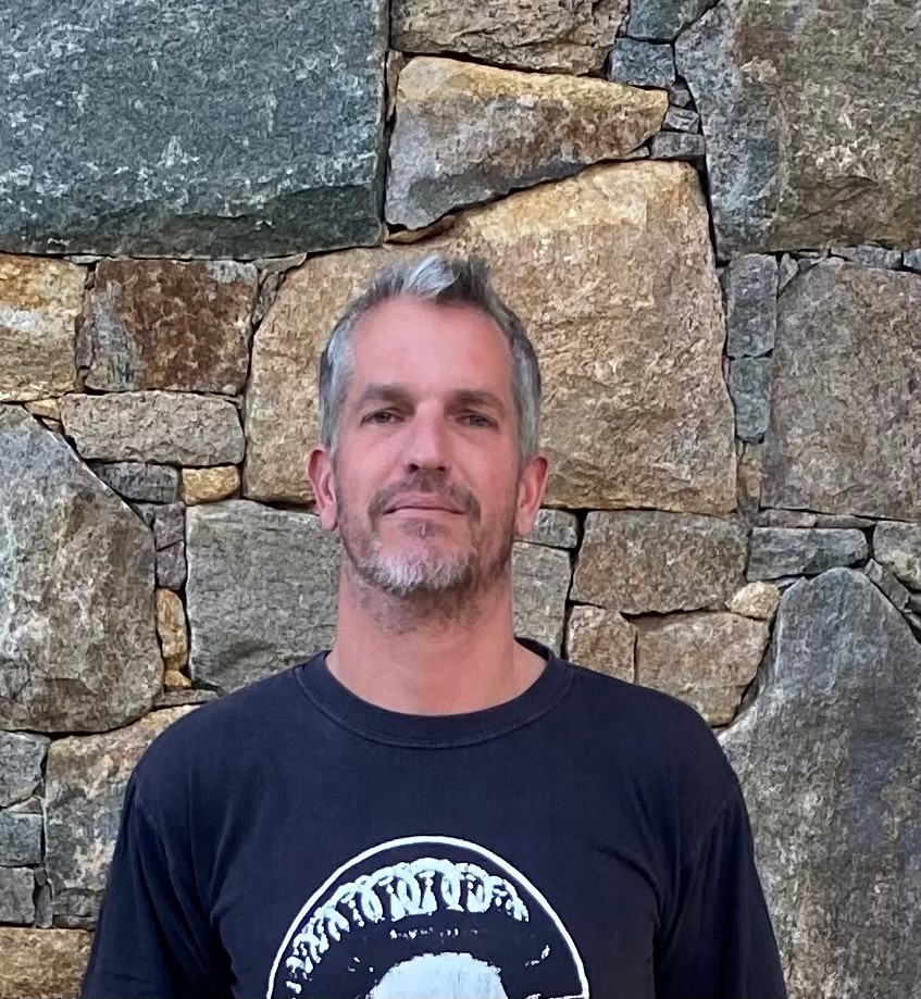 A man with short salt and pepper coloured hair and slight beard stands in front of a stone wall 