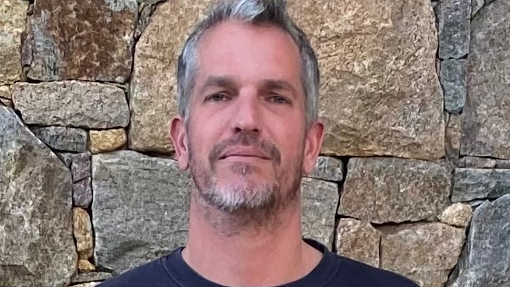 A man with short salt and pepper coloured hair and slight beard stands in front of a stone wall 