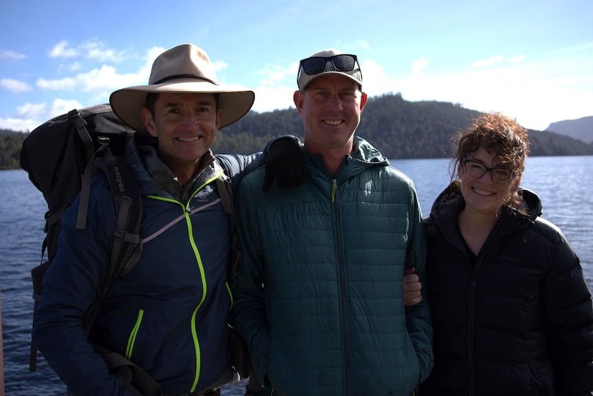 Two men and one woman dressed in hiking gear and hats stood on the end of a pier with a lake vista in the background. 