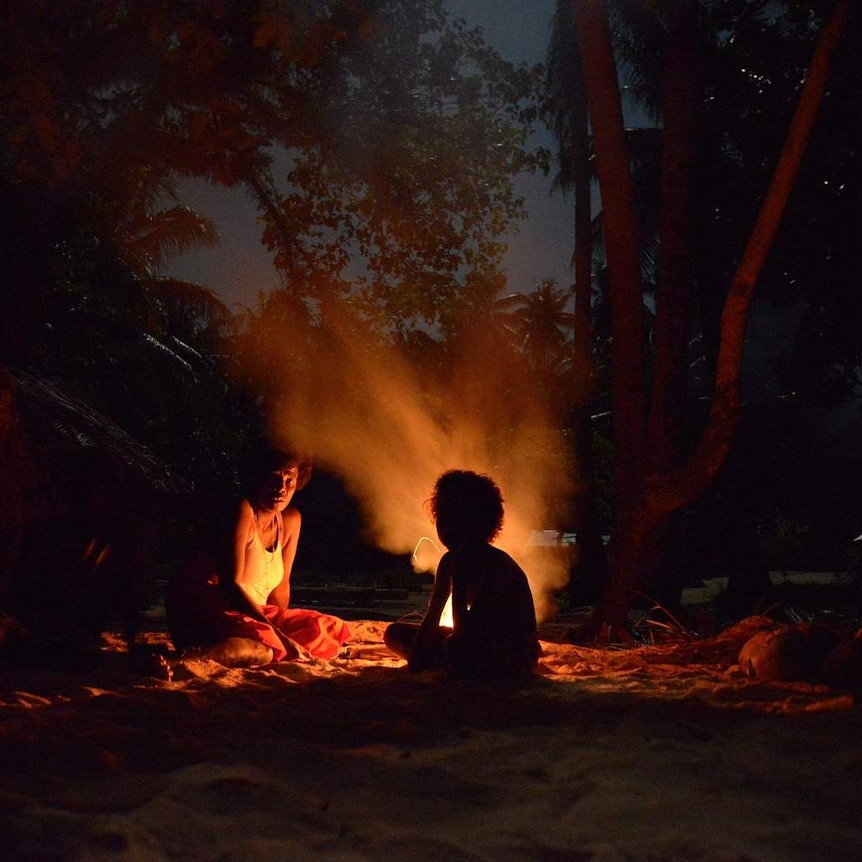 Two people sit in the sand by the fire beneath trees.