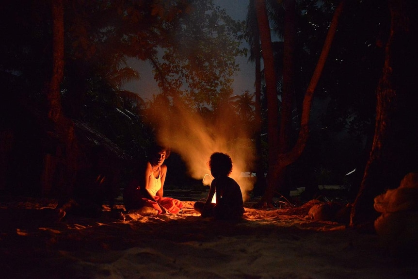 Two people sit in the sand by the fire beneath trees.