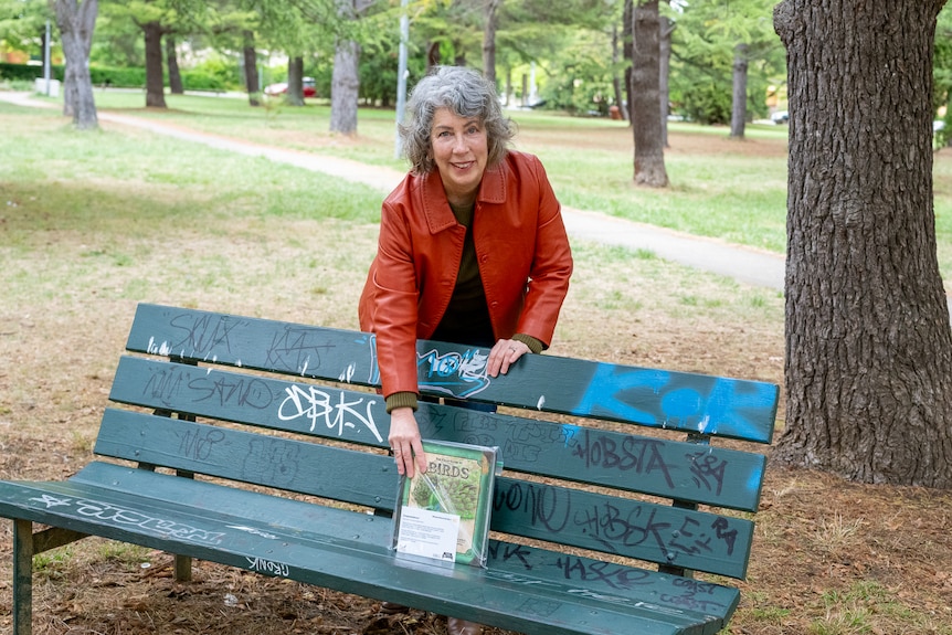Woman wearing an orange jacket places a book onto a park bench. 