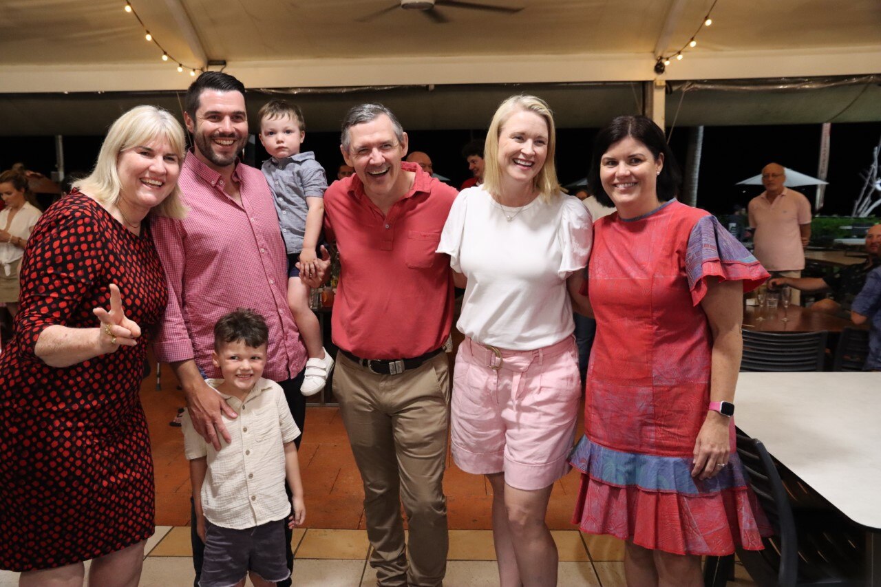 Five adults smiling and posing for a camera, wearing shades of red. One of them holds two small children.