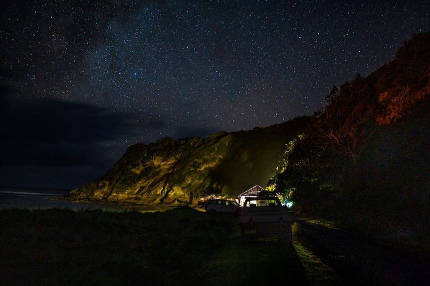 The night sky full of stars can be seen from a distance, as can a small gathering of people