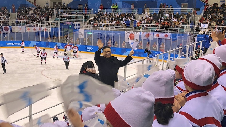 A man waves a flag in front of a stand full of cheerleaders in an ice hockey arena.