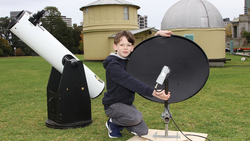 An 11-year-old boy beside a black satellite dish and a white telescope.