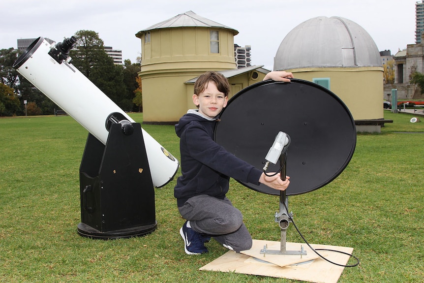 An 11-year-old boy beside a black satellite dish and a white telescope.