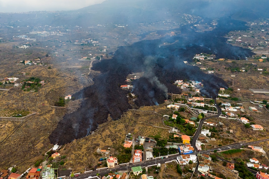 An aerial image of black lava spreading through green bushland, nearby is a cluster of homes