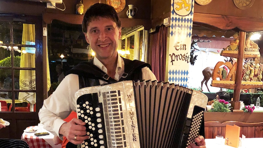 A smiling man in a white shirt and black waistcoat plays the accordion in a German-themed restaurant.
