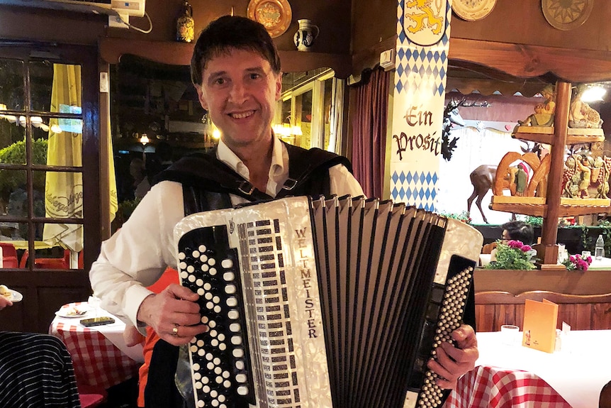 A smiling man in a white shirt and black waistcoat plays the accordion in a German-themed restaurant.