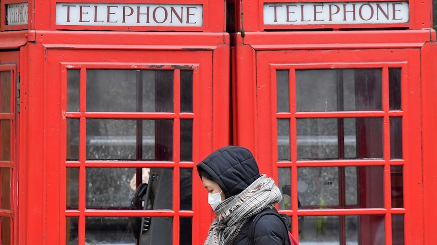 A woman in winter clothes and with a face mask looks at her phone as she walks past red phone boxes