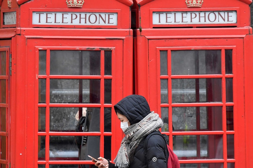 A woman in winter clothes and with a face mask looks at her phone as she walks past red phone boxes