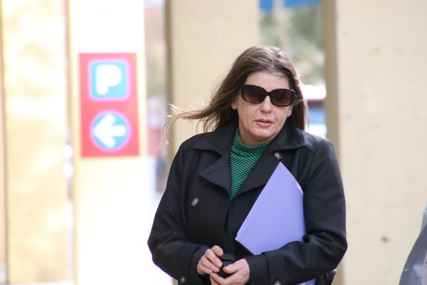 A woman wearing sunglasses and a jacket walks with a folder tucked under her arm.