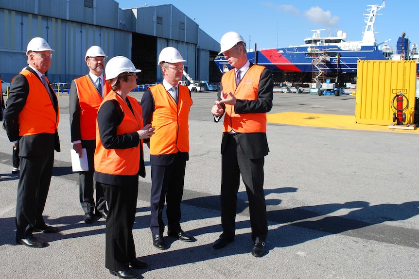 Five people in suits and hi-vis vests stand talking at a shipbuilding yard in Henderson.