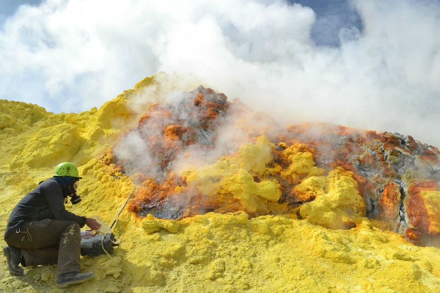 A researchers in gas mask samples gas coming from bright yellow rock face.