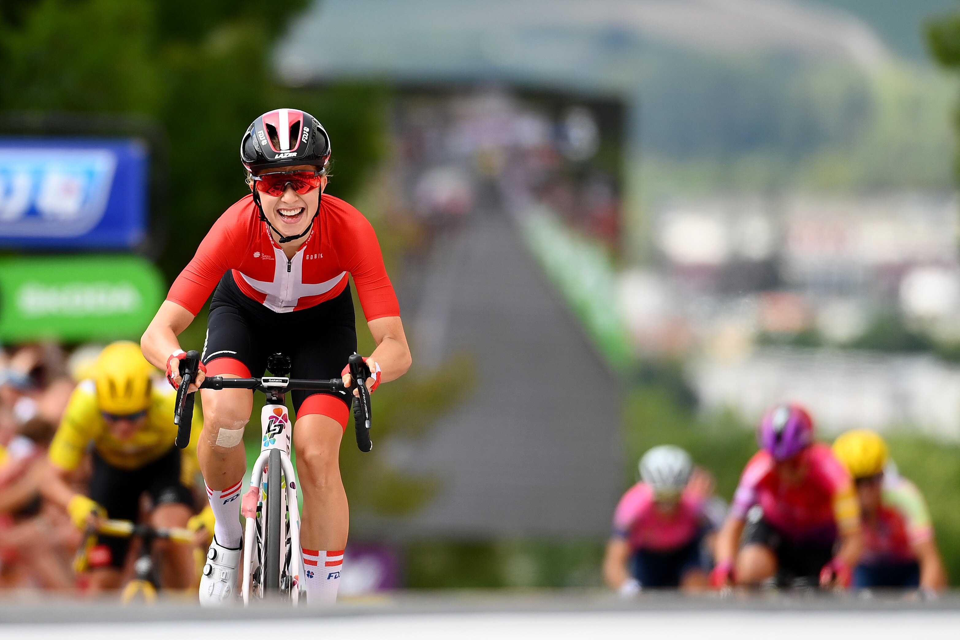 A cyclist dressed in red grins as she wins a Tour de France Femmes stage, with the yellow jersey behind her.