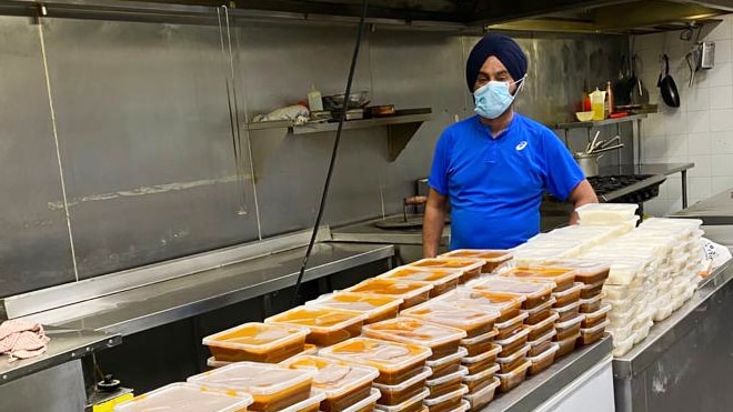 Ravinder Sahni standing in a commercial kitchen. Cooked vegetarian meals are in plastic containers on the table.