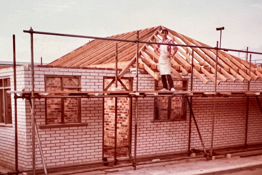 An old sepia image of a woman in an apron standing on a ladder by a house being built.