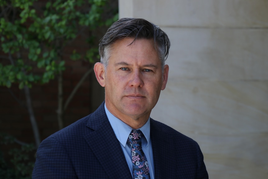 Brad Pettitt, wearing a dark suit, blue shirt and tie, looks at the camera. Behind him is a wall and a tree.