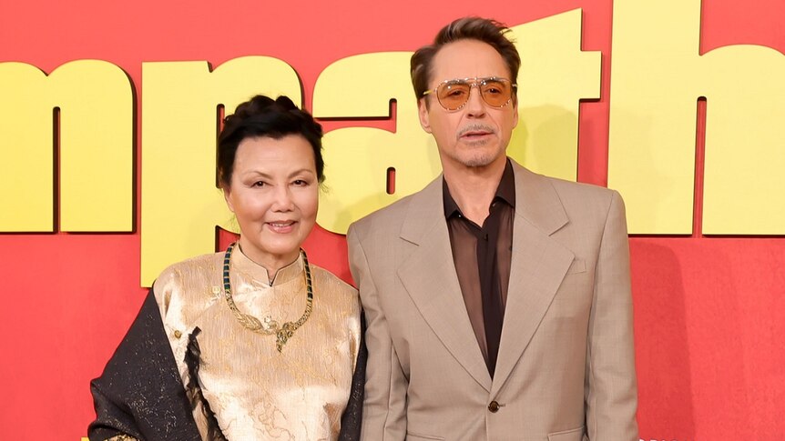 A Vietnamese woman and a middle-aged man wearing glasses pose against a red background at a film premiere