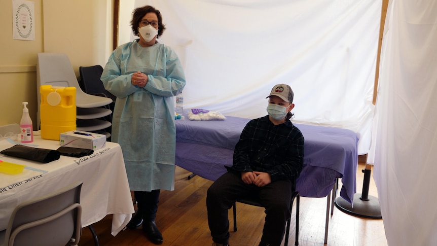 Cash Reed sits in a chair with a nurse dressed in PPE by his side. The two wear masks. Vaccination supplies visible as well.