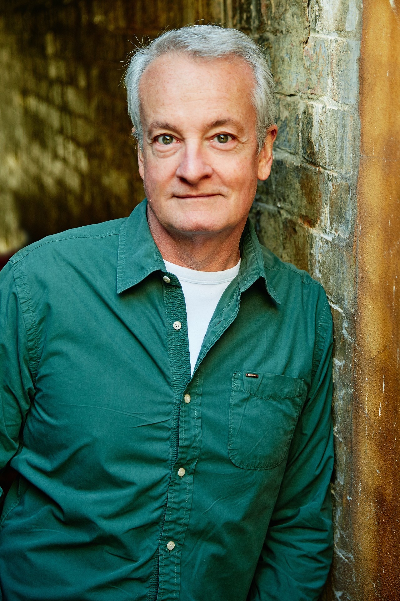 A 60-something man looks into the camera. He's leaning against a brick wall, while wearing a dark green button-up shirt 