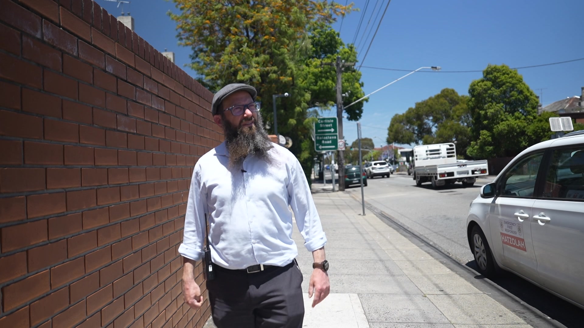 Um homem com uma longa barba, óculos e um chapéu cinza com uma camisa de colarinho branco olha para fora da câmera ao lado de uma cerca de tijolos laranja.