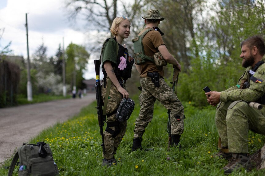 A woman in military uniform stands on a grassy roadside next to two male soldiers. 