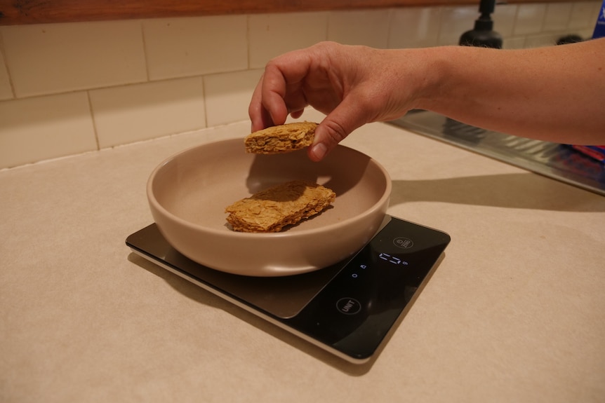 A hand puts a wheat breakfast cereal into a light bowl on a black set of scales