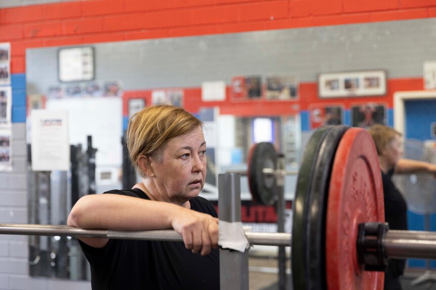 A middle-aged woman leans on a weightlifting bar, wearing a serious expression.