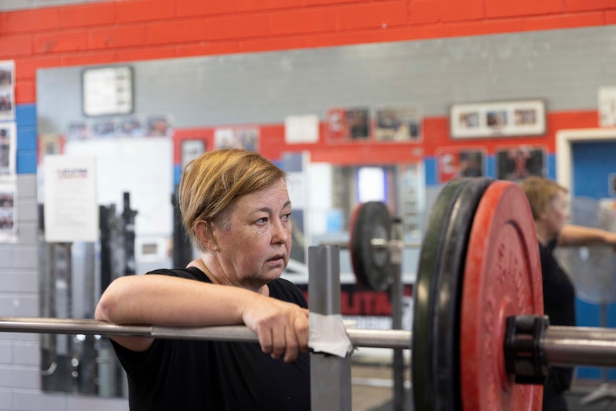 A middle-aged woman leans on a weightlifting bar, wearing a serious expression.