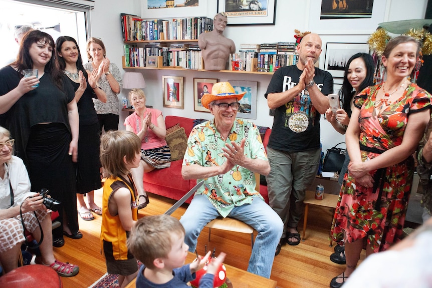 An older man wearing a cowboy hat while clapping surrounded by family members in a living room