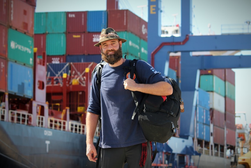A man carrying a backpack stands in front of a large cargo ship docked at a port