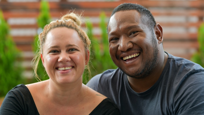 Tara and Luke Kaspar look directly at the camera and laugh. They are in their backyard, and there are some greenery behind.