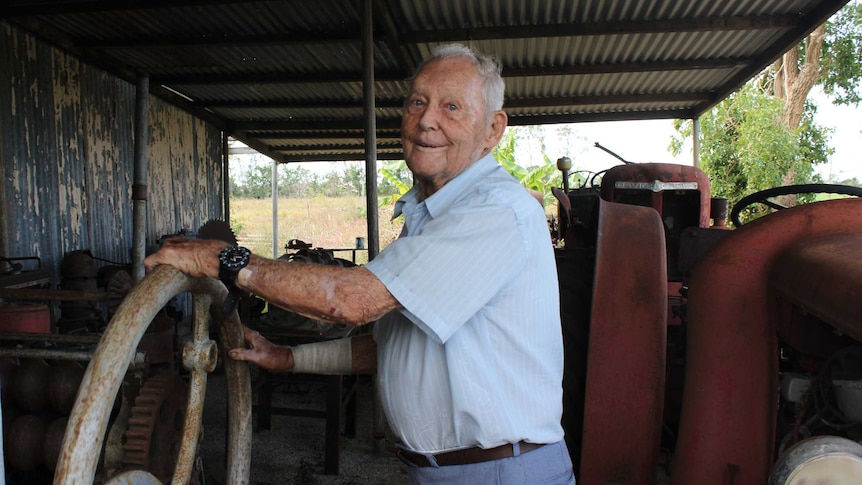 A man with his hands on an old rusty steering wheel smiling in front of old farming equipment