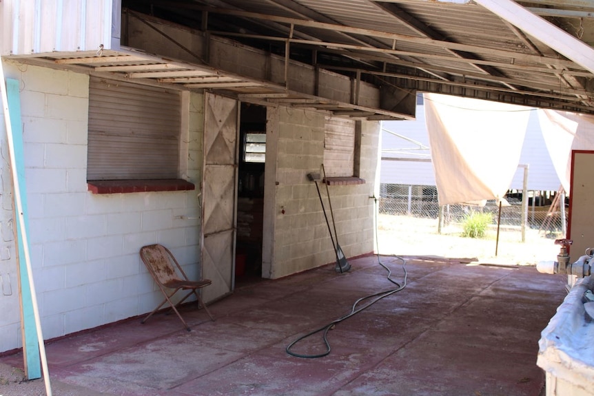 Un área de concreto afuera de una casa de bloques, una silla oxidada apoyada contra una pared, ropa tendida al sol.