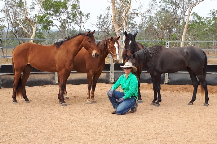 Rebecca kneels in a green shirt in front of four horses.