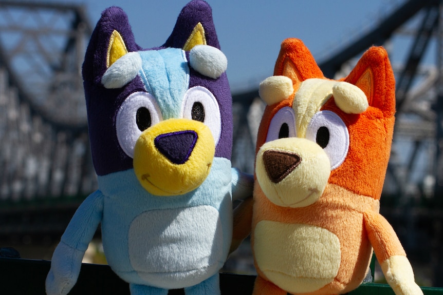 Plush toys of Bluey and Bingo in front of a bridge