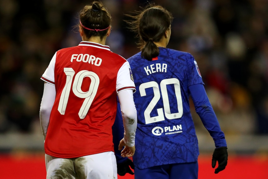Two soccer players, one wearing red and white and the other wearing blue, have their backs to the camera showing their numbers