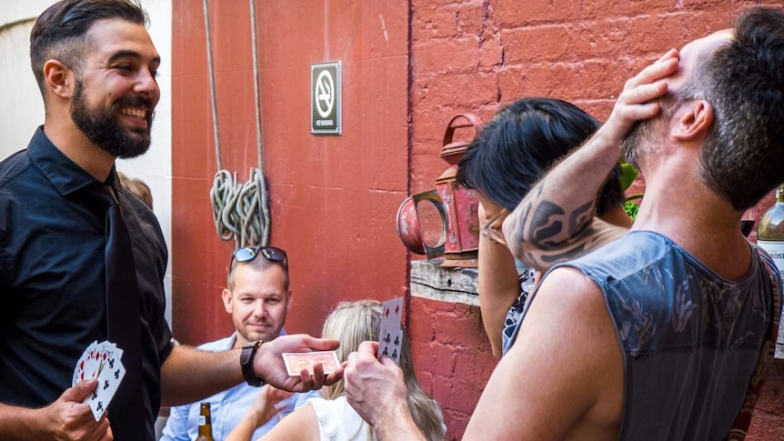 A young bearded man holds a card in his hand while a man and woman shriek with laughter.