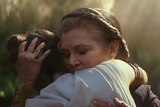 Wearing crown braid and with eyes closed Carrie Fisher embraces Daisy Ridley, both illuminated by golden light in forest.