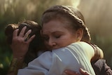 Wearing crown braid and with eyes closed Carrie Fisher embraces Daisy Ridley, both illuminated by golden light in forest.