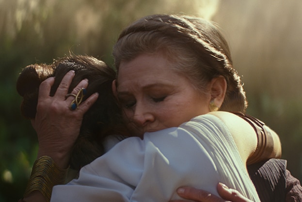 Wearing crown braid and with eyes closed Carrie Fisher embraces Daisy Ridley, both illuminated by golden light in forest.