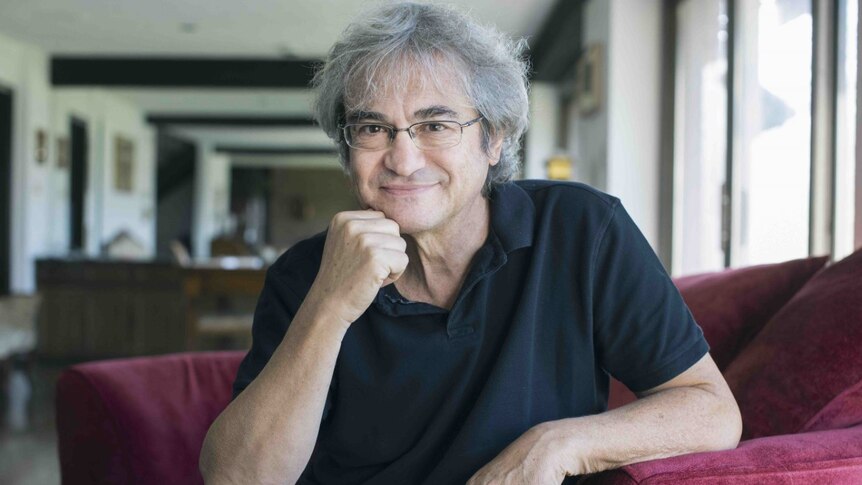 Carlo Rovelli with silver wavy hair sitting on a red couch in a black short sleeved shirt and glasses smiling at the camera