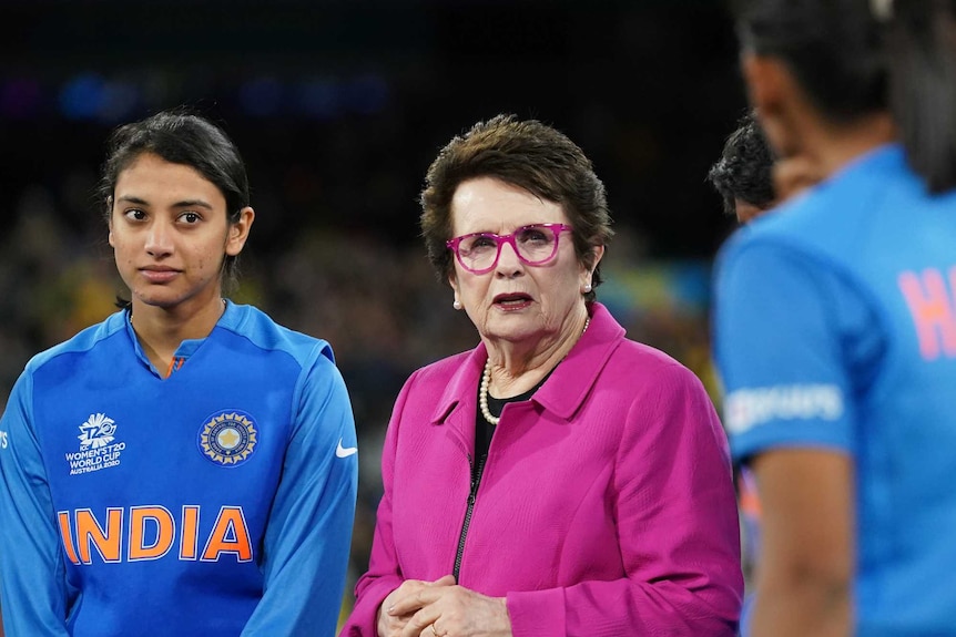 Billie Jean King looks to the camera surrounded by players from India wearing blue tops. She wears fuchsia top and glasses