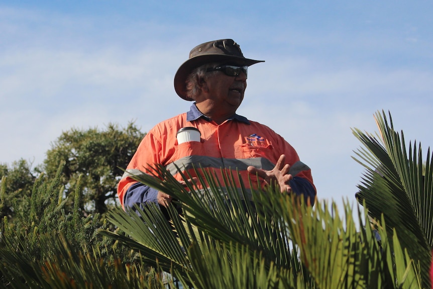 A man standing in bush land wearing a bright work shirt and a hat. 
