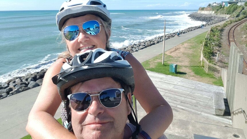 Tina and Steve Dixson smile at the camera while wearing bike helmets and standing next to a linear path near the sea.