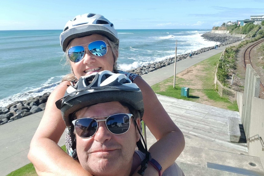 Tina and Steve Dixson smile at the camera while wearing bike helmets and standing next to a linear path near the sea.